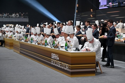 Kucharze biorący udział w finale siedzą przy długim stole, w tle telebim